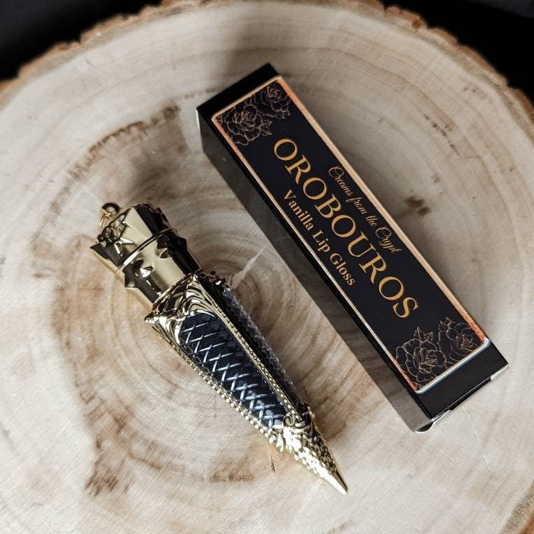 A luxurious gold bottle of OROBOUROS black lip gloss with a French vanilla design, displayed next to its packaging on a wooden surface.