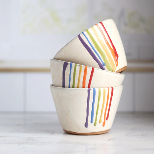 "Stack of three cream colored ceramic bowls with rainbow drip glaze patterns, artistically arranged showing the colorful striped designs"